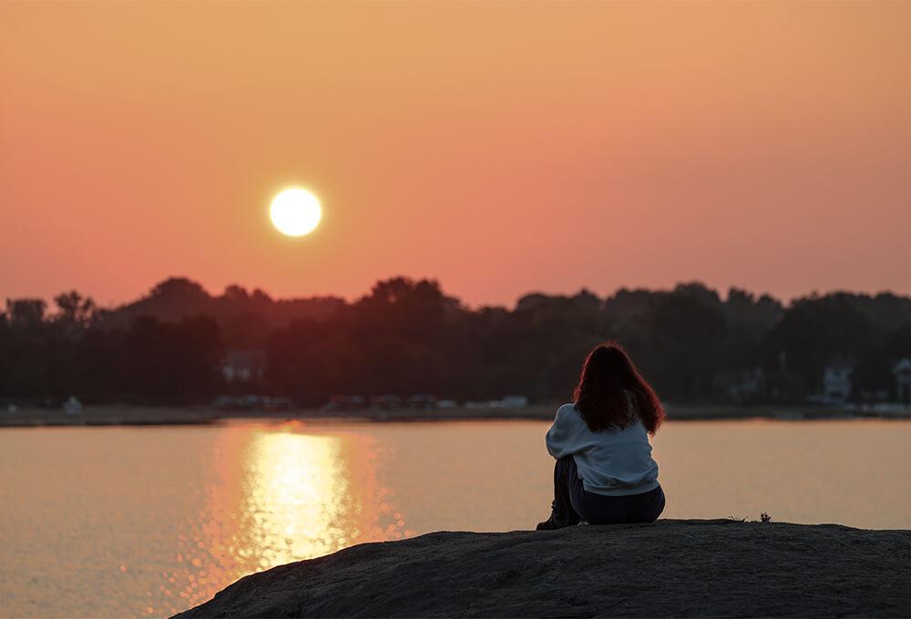 A woman sitting quietly on a wooden dock at sunrise, looking across a calm lake