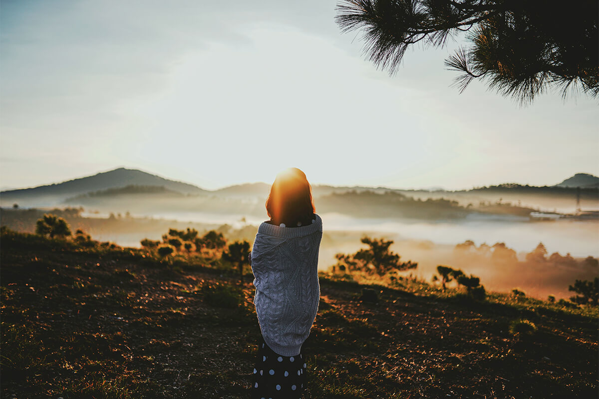 Woman sitting quietly on a hillside at sunrise, reflecting on faith and seeking a deeper connection with God.