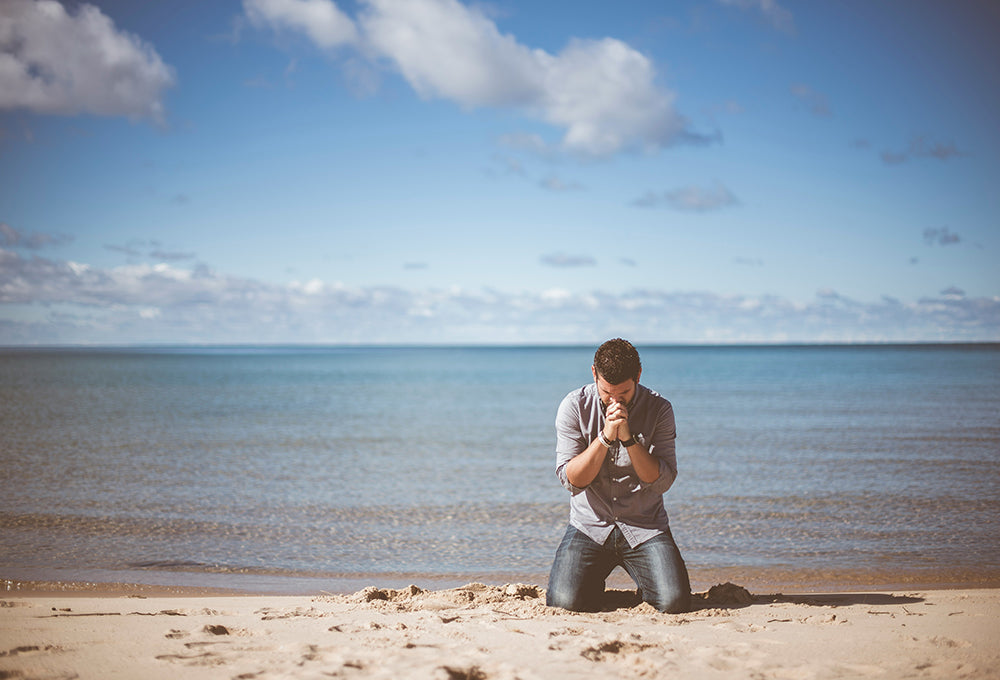 Middle aged man praying on the beach next to a calm ocean