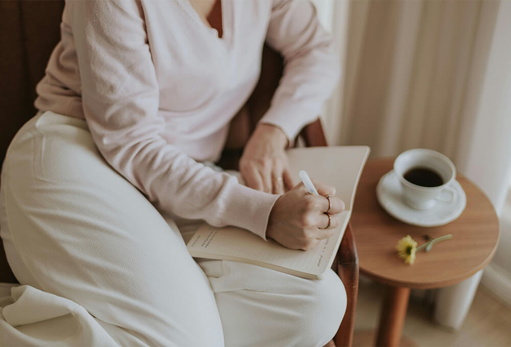 Woman writing gratitude prayers in her journal with coffee nearby