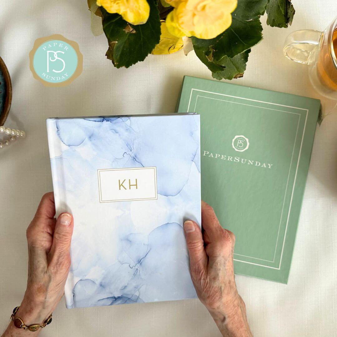 Older woman holding Sky personalized scripture journal by Jenn Thatcher over a white linen table with flowers and pearls, showcasing a calm reflective setting with a unique curated set of Bible verses