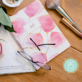 Hardcover Blushing Floral Bible journal by Elizabeth Lalama styled on a wooden desk with reading glasses and makeup, open journal displaying personalized scripture pages for quiet time and journaling