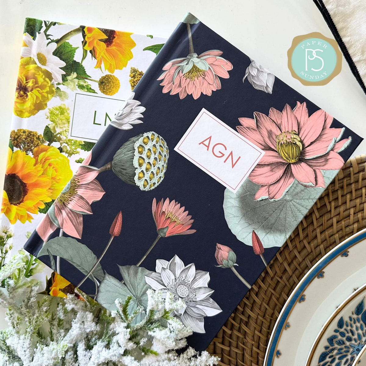 Water Lily and Sunflower Cross personalized scripture journals stacked together beside a classic dinner plate setting, showcasing floral designs and curated Bible verse journals for meaningful daily use and gifting